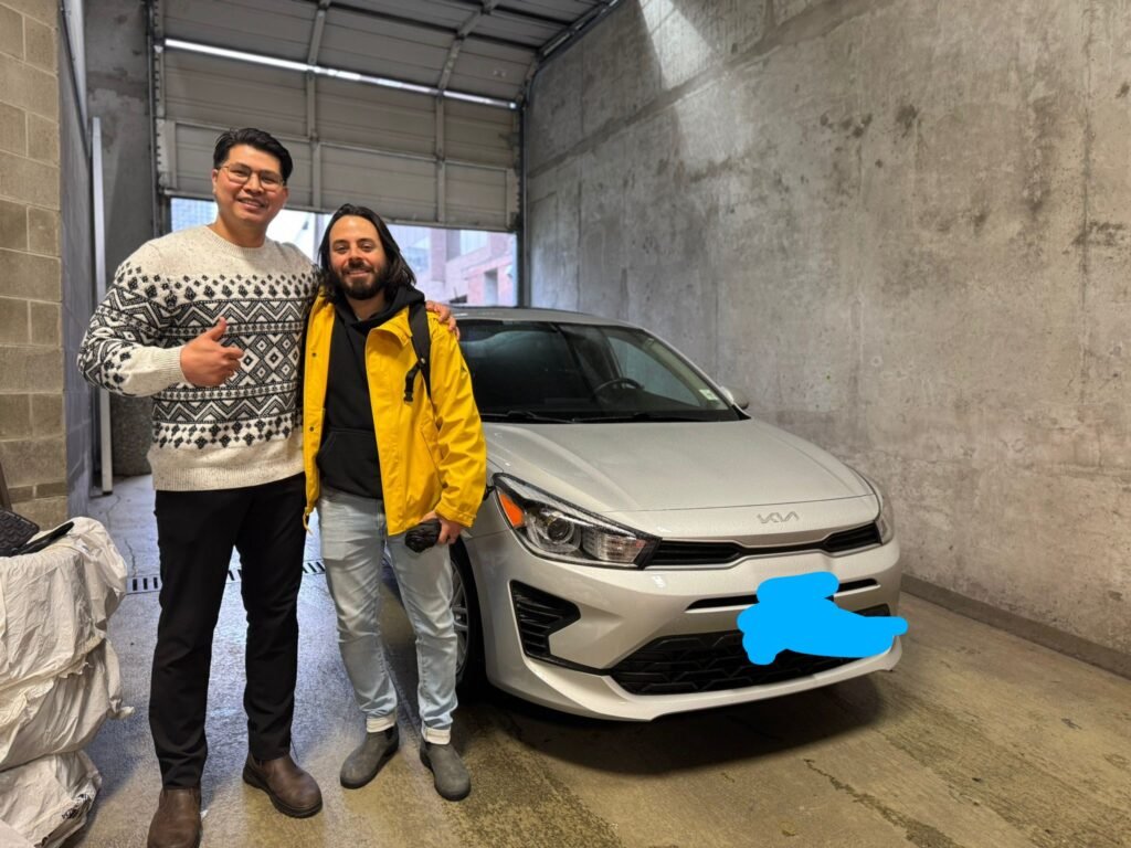 Ken from GettingDeal.com stands beside a smiling customer and his new white Kia in an industrial garage setting, celebrating a successful car purchase.