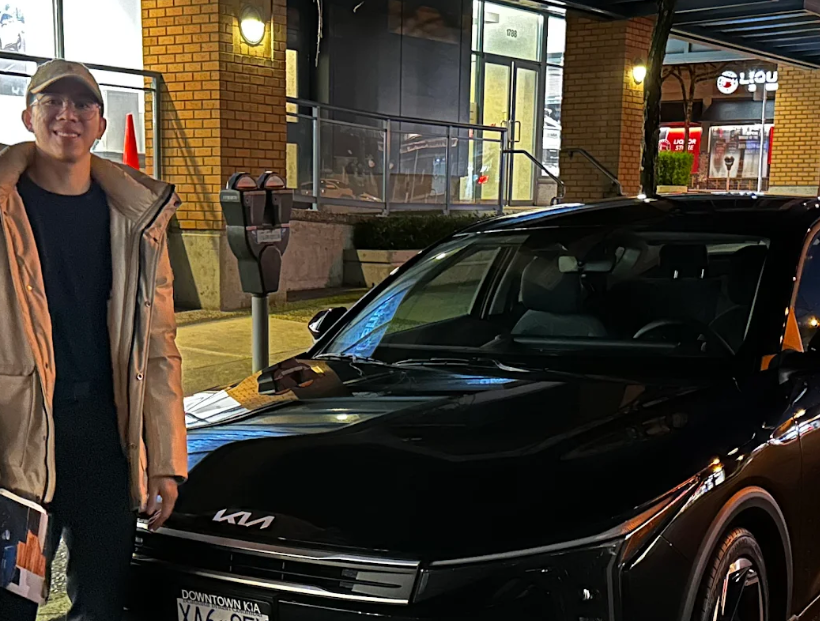 A young woman smiling beside a black Kia EV parked on a downtown street at night.