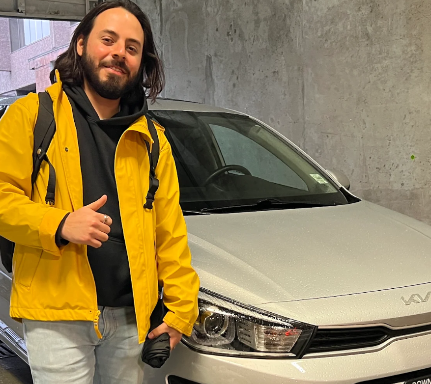 A smiling man in a yellow jacket giving a thumbs-up next to his white Kia sedan in an urban parking area.