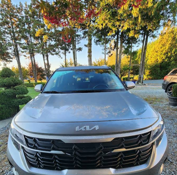Front view of grey Kia SUV parked in a driveway with autumn trees