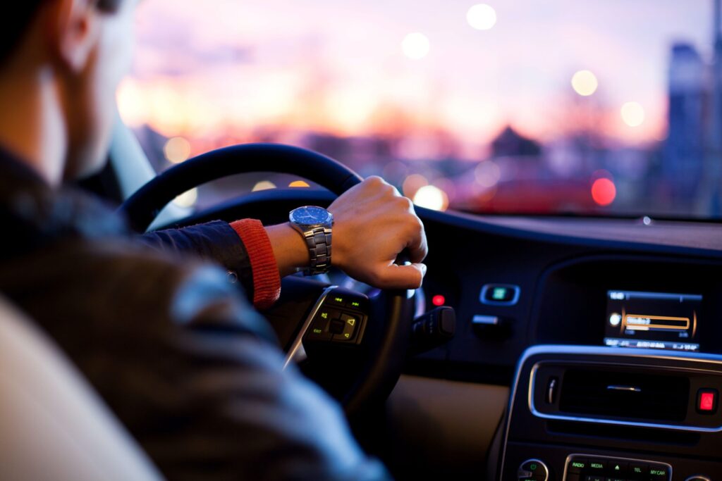 Driver's hand on the steering wheel with city lights in the background during sunset