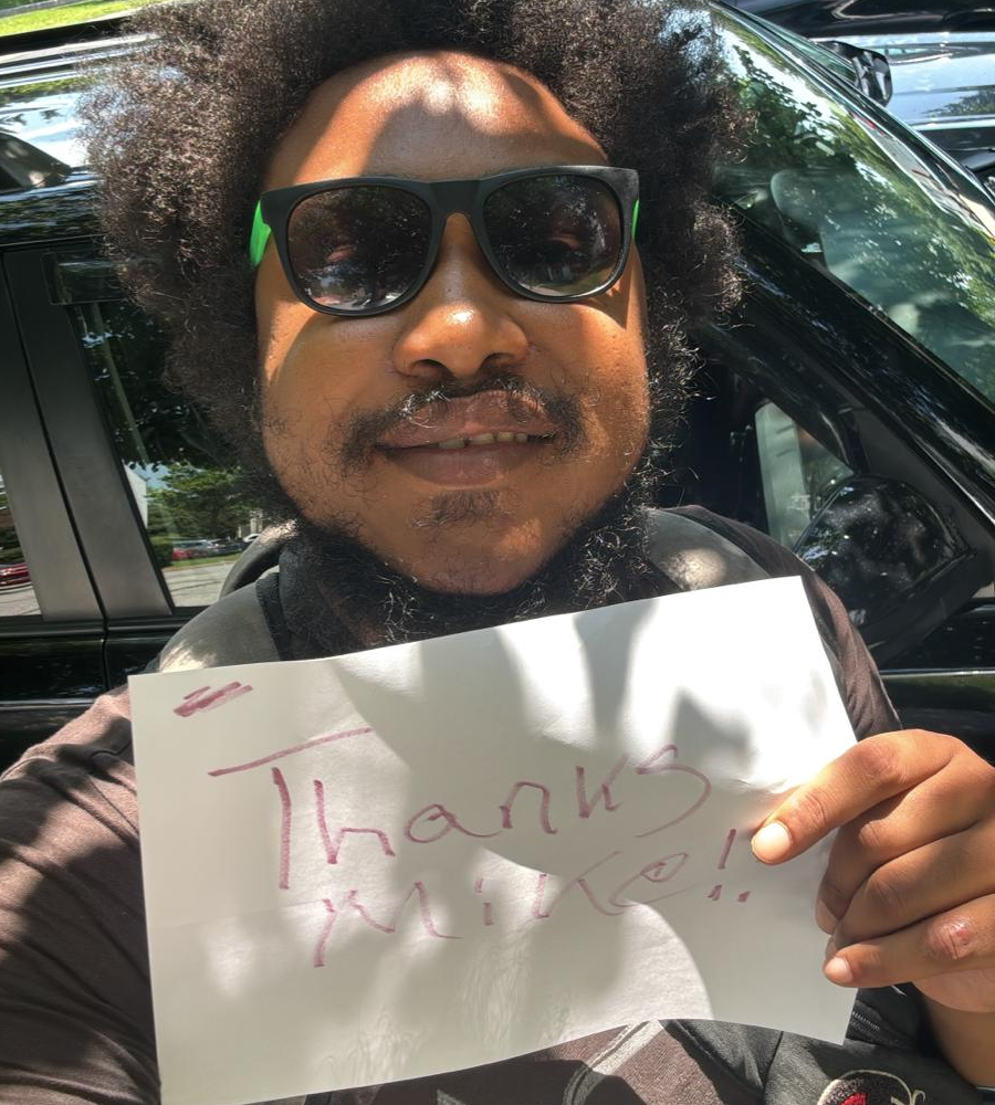 Smiling man holding handwritten "Thanks Mike!" sign in front of a black car, wearing sunglasses on a sunny day.