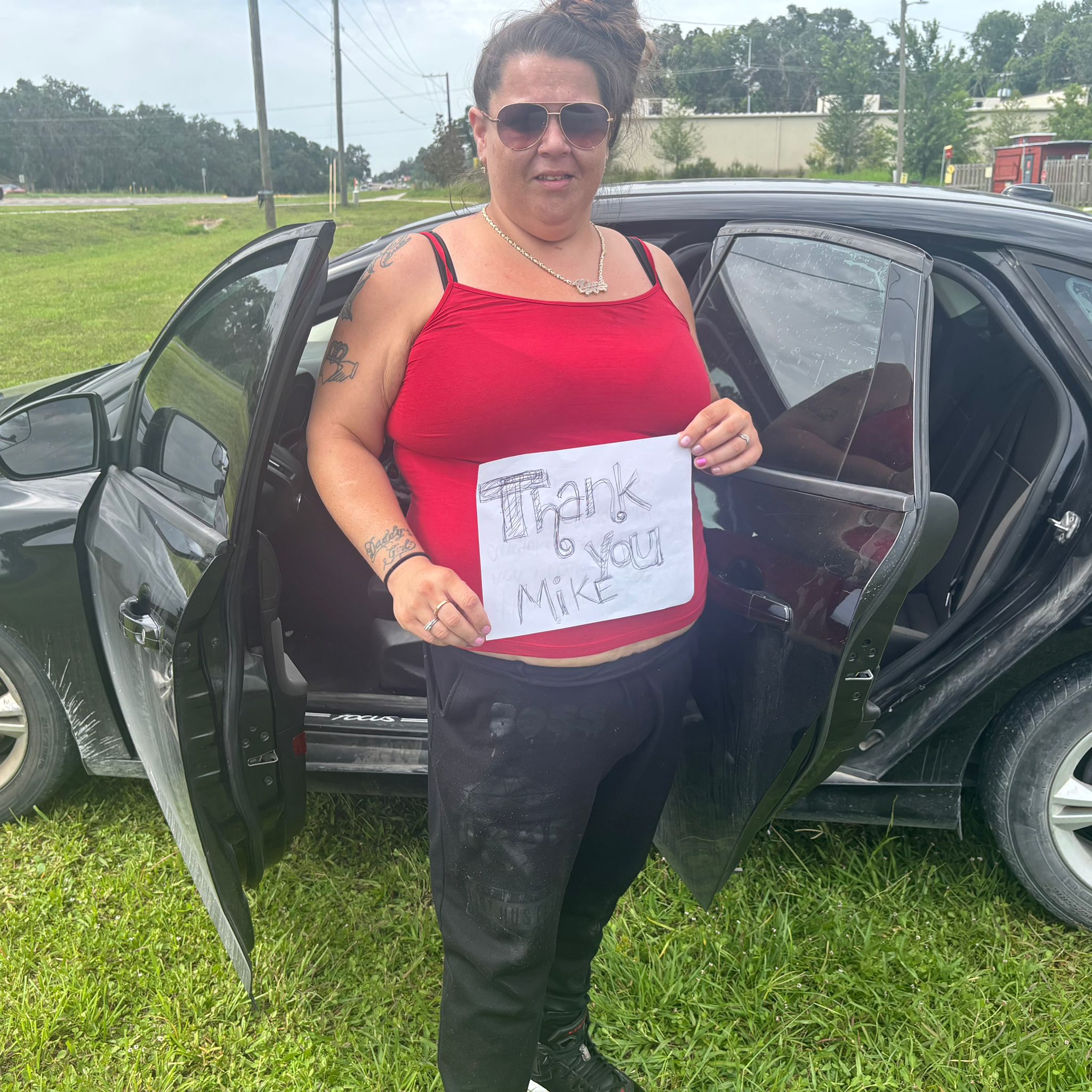 A woman in a red tank top and sunglasses stands beside her black car, holding a handwritten “Thank you Mike” sign, smiling after getting approved through GettingDeal.com.
