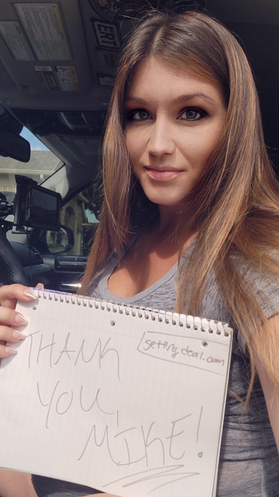 Smiling woman sitting in her car holding a handwritten “Thank You Mike! GettingDeal.com” sign, showing appreciation after getting approved for a car loan.